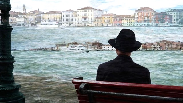 someone in a dark hat sits on a park bench, facing away from us, looking at the urban skyline across a river; in the water is a miniature version of the same skyline