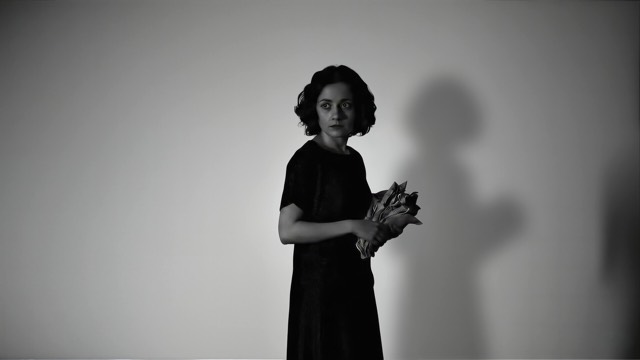 against a plain white backdrop, we see a young woman with medium length dark hair holding some crumpled paper that looks a bit like a bouquet of flowers