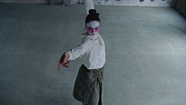 a woman stretches her right arm toward the camera; her face is painted white with large pink shapes around both eyes and cheeks