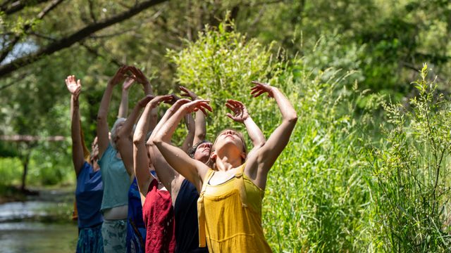 six dancers wearing bright solid colors stand in a shallow creek, chests open and arms raised to the sky