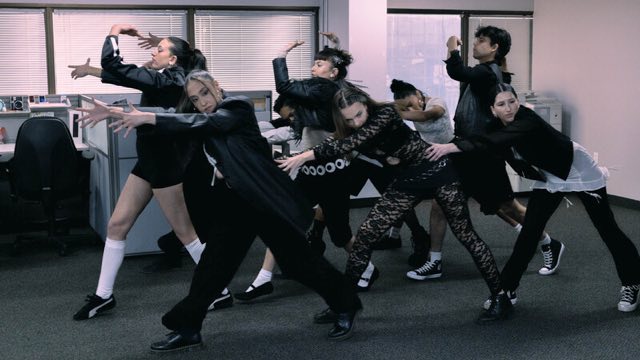 nine young dancers in an office building, all wearing black and/or white, lean forward in various poses