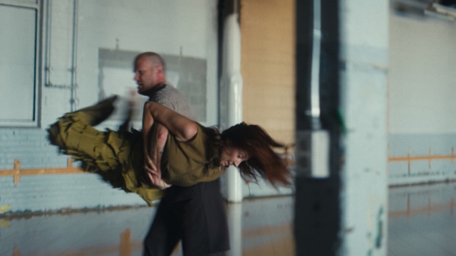 in a large empty industrial area with a polished floor, a male dancer with a shaved head lifts and spins his partner by the waist