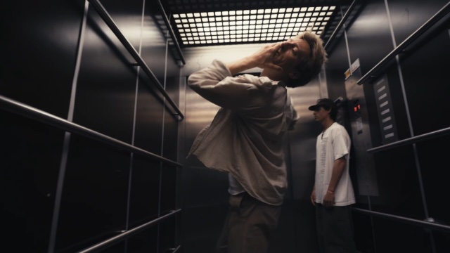 two men in a black elevator; the one in the foreground tilts his head back - left hand on face - while the one in the background stands motionless