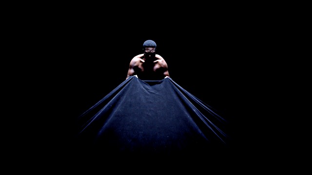in a pitch black room, a dark skinned man with short black hair pulls on a black velvet curtain