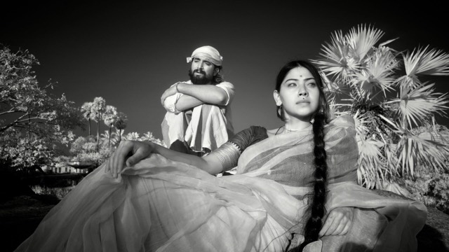 a monochrome photo of a woman with a long dark braid looking off frame to her left while a man with a dark beard and mustache sits behind her, arms folded over knees