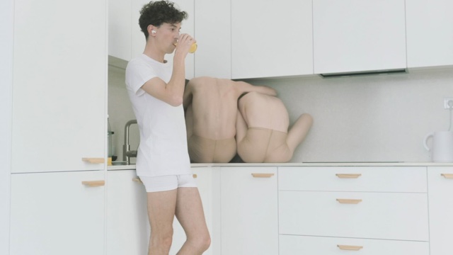 in a sparse white kitchen, a young man in a white tee shirt and white underwear drinks a glass of orange juice while behind him, two shirtless people sit hunched over on the counter