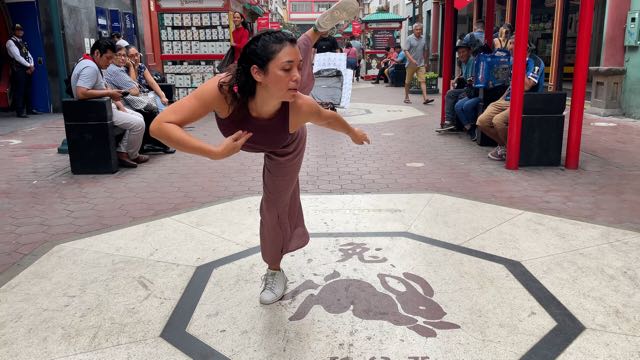 in a public street with seats and shops in the background, a woman wearing mauve colored clothing stands on her left leg, leaning fully forward