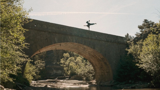 in the distance, a dancer stands on one hand atop a stone bridge over a river
