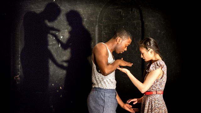 a dark skinned man and tan skinned woman use their hands to interact playfully; their shadows are cast on the wall behind them