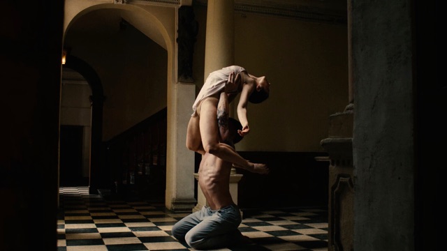 from above, a shirtless man kneels on a black and white checkered floor, lifting a woman in a tee shirt and no pants by her shoulder blades