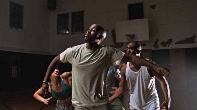 in a rundown basketball gym, a handful of dark skinned dancers move with arms loose