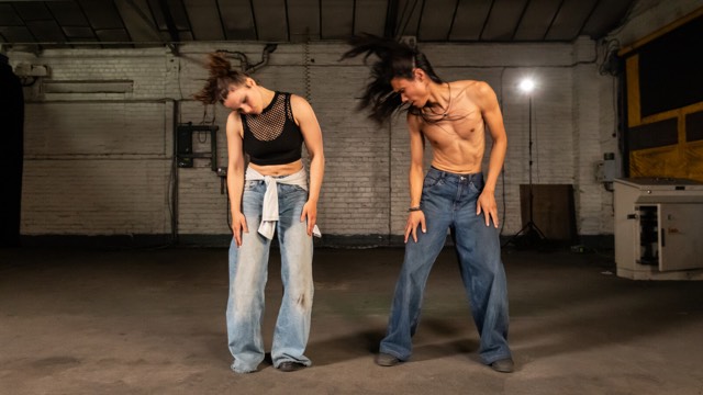 a male and female dancer - each wearing blue jeans - swing their heads to their right, letting their hair flail