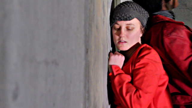 a woman in a bright red jacket lays on a concrete floor or road with her right cheek pressed to it; the image is rotated 90 degrees such that she appears vertical