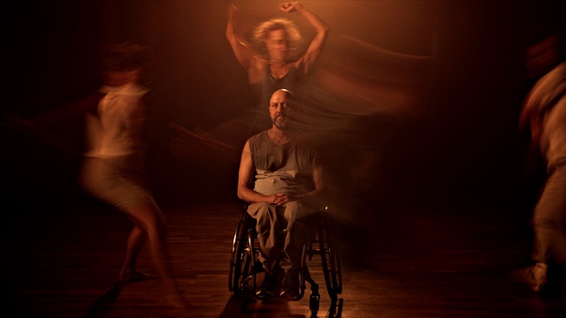 in a room lit dimly by warm light, a man in a wheelchair faces the camera with an inscrutable expression; around him are the motion-blurred forms of three dancers
