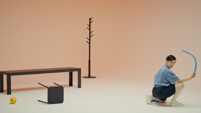 in a sparsely decorated room where a cream colored floor gives way seamlessly to peach colored walls, a dancer in shorts and a baggy short sleeved shirt kneels and faces the camera