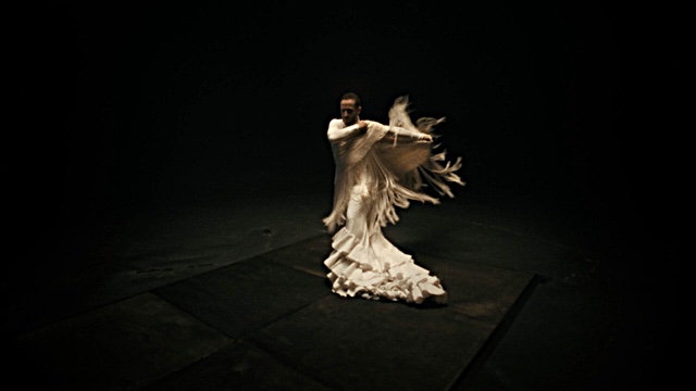 a man in a long, flowing cream colored dress and shawl dances through a black room