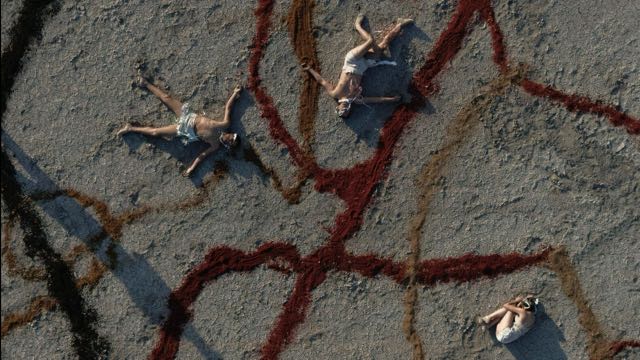 from overhead, we see three dancers sprawled out on a sandy plain streaked with thick lines of maroon and brown