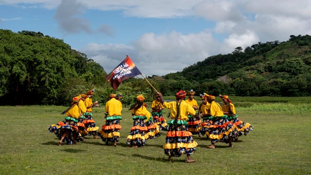 in a grassy field surrounded by lush hills, a dozen dancers in yellow shirts and long dresses moves in a circular formation; one is holding a flag with indiscernible writing