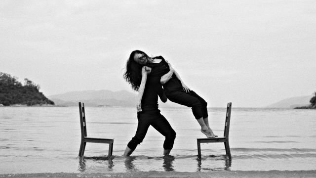 two dining room chairs face each other in the shallow water of a large body of water; on one, a woman stands, leaning off while another dancer supports her