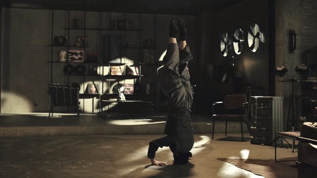 a dancer stands on their head, palms on the floor and facing away from the camera in front of a wall shelf filled with intriguing objects