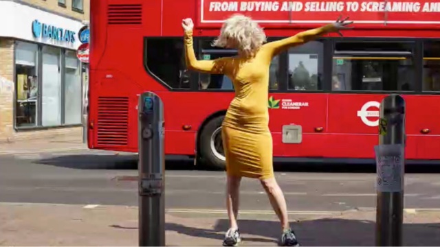 a woman in a tight mustard colored knee length dress and tennis shoes stands on a sidewalk with a bright red city bus behind her; her wild blond hair obscures her face