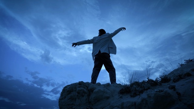 from below, a dancer in dark dress pants and a white suit jacket faces upward toward a dark blue sky