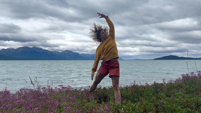 in a field of purple flowers on the shore of a large body of water, a woman in a yellow sweatshirt and pink shorts lifts her left hand overhead