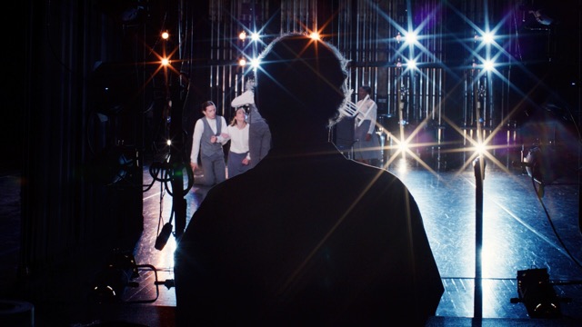 we see a well lit stage with performers in grey and white formal wear, but the scene is obscured somewhat by the silhouette of a person in the foreground