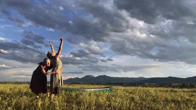 in a grassy field with mountains in the background, one older woman reaches her hands up to the cloudy afternoon sky while another holds her gently by the waist