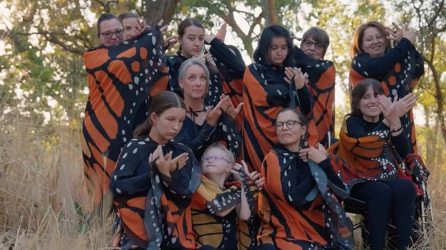 eleven people spanning the age spectrum make butterfly shapes with their hands while wearing monarch butterfly-patterned capes