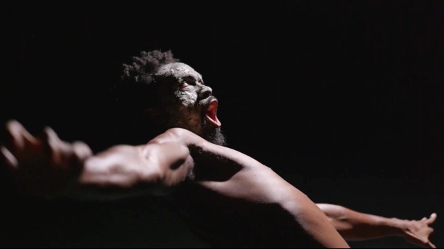 against a black background, a shirtless dark skinned man with dark face paint leans backward, mouth open as though singing