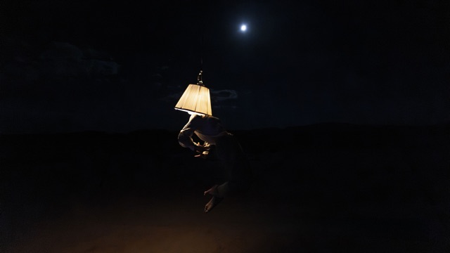 a table lamp vaguely illuminates a person holding it in an otherwise pitch dark room