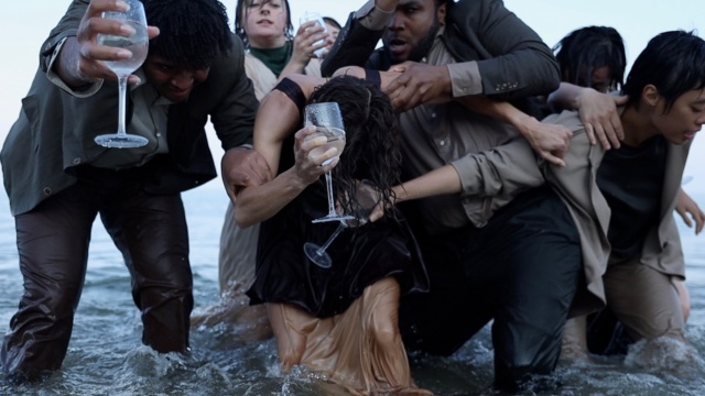 several young dancers in formal attire trudge through knee-deep water, holding wine glasses