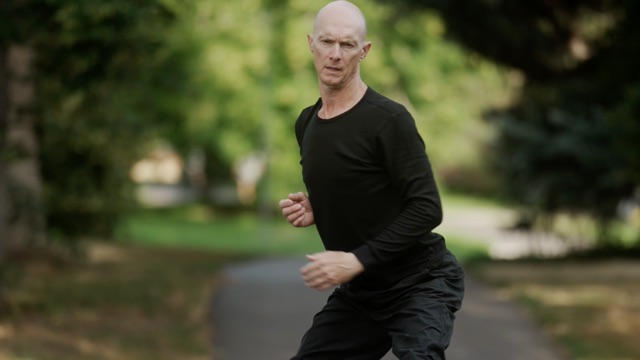 a lean, light skinned man with a shaved head, wearing all black, dances with hips out in a park