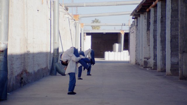 in an concrete industrial alley, three pairs of dancers in white shirts and blue pants do partner lifts