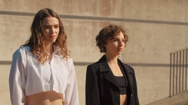two young light skinned, brown haired women - one wearing a white top and one wearing a black top and jacket - look with determined expressions off the frame