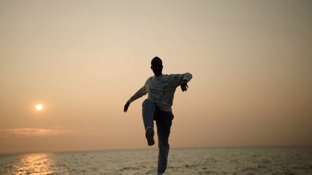 with the backdrop of the sun setting over the ocean, a dark skinned man lifts his right knee with arms out and elbows bent