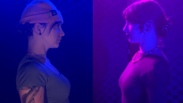 in a dark room illuminated by reddish light, two differently dressed versions of the same light skinned woman face each other