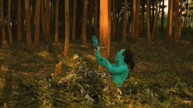 in a grove of similar looking trees, a man painted turquoise emerges from a pile of leaves on the ground