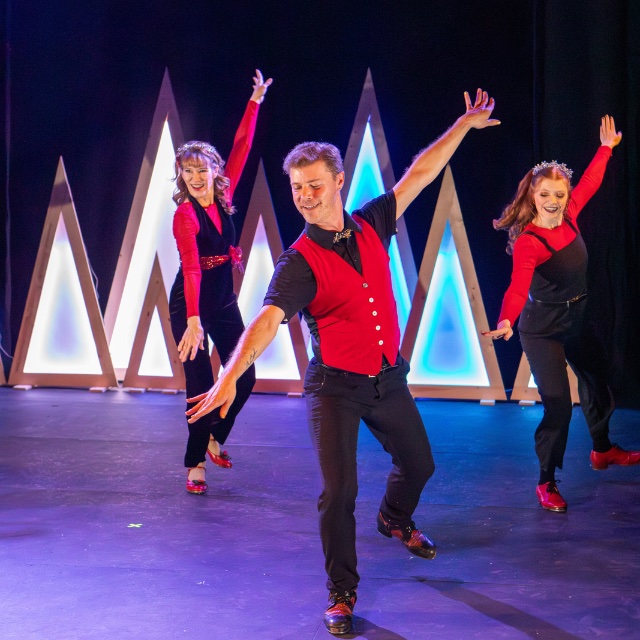 three tap dancers in red and black costumes tap in unison on a stage with brightly lit triangles at the back